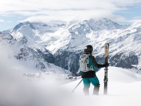 Ein Mann in Skibekleidung und Helm steht im Pulverski mit Scott Freeride Ski in der Hand und Blickt in die verschneite Berglandschaft