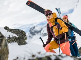 Zwei Frauen gehen eine Tour einen Berg hinauf, die Ski sind am Rucksack befestigt