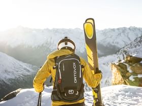 Ein Mann in Skibekleidung und Helm mit Ortovox Rucksack steht mit Kästle Ski am Bergrand und schaut in die winterliche Berglandschaft