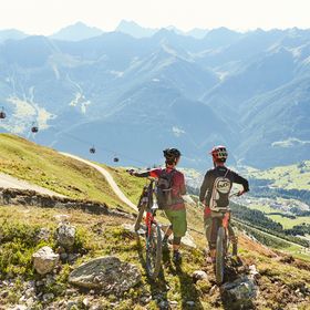 Zwei Biker stehen mit ihren Mountainbikes an einem Biketrail und blicken auf die Seilbahn und Berglandschaft von Serfaus-Fiss-Ladis
