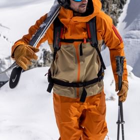 Ein Mann läuft am Grat eines verschneiten Gipfels entlang und trägt seine Ski auf der Schulter