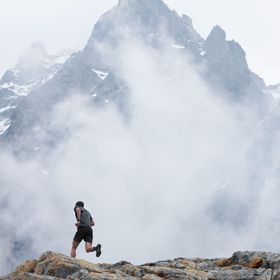 Ein Mann läuft in On Bekleidung und Schuhen auf einem Grat einen Berg hoch. Im Hintergrund ist ein hoher Berggipfel hinter Wolken