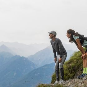 Eine Frau und ein Mann in On Running Bekleidung und Schuhen stehen auf einem Steig in der Bergwelt von Serfaus und blicken in die Landschaft