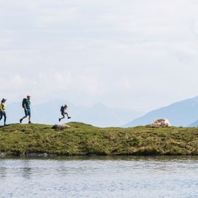 Eine junge Familie geht am Högsee Ufer spazieren. Der Junge springt hoch von einem Stein. Im Hintergrund ist eine Berglandschaft 