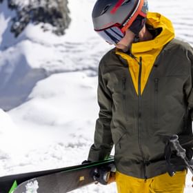 Ein Mann läuft am Grat eines verschneiten Gipfels entlang und trägt seine Ski in der einen einen, die Stöcke in der anderen Hand