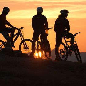 Eine Gruppe von Bikern schaut auf einem Trail den Sonnenuntergang in den Bergen zu