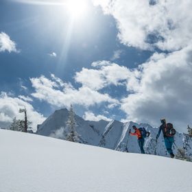 Eine Gruppe von Skifahrern geht eine Tour einen Berggrat hinauf, die Ski sind am Rucksack befestigt im Hintergrund ist eine Bergpanorama mit verschneiten Gipfeln