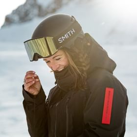Eine Frau lächelt nach einer Abfahrt im Pulverschnee in den Bergen in einer Mountain Studio Jacke und Helm und Brille zufrieden in die Kamera