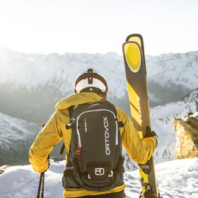 Ein Mann in Skibekleidung und Helm mit Ortovox Rucksack steht mit Kästle Ski am Bergrand und schaut in die winterliche Berglandschaft