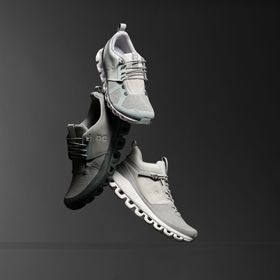 verschiedene Schuhmodelle der Marke On in der Farbe Weiss und Grau vor einem grau schwarzen Hintergrund
