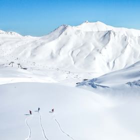 Eine Gruppe von Snowboardern fährt durch den Tiefschnee im Masnergebiet Serfaus. Im Hintergrund sind weiß verschneite Berge und die Masnerhütte