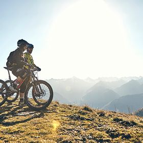 Zwei Biker stehen am Rande eines Trails und blicken auf ihren Mountainbikes sitzend in die Berglandschaft von Serfaus-Fiss-Ladis