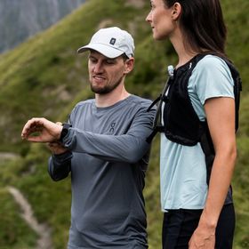 Eine Frau und ein Mann in On Running Bekleidung und Schuhen stehen auf einem Steig in der Bergwelt von Serfaus und checken die Zeit auf einer Smartwatch