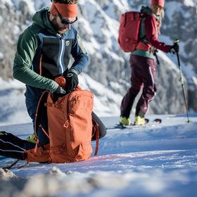 Ein Mann packt in einer weiß verschneiten Berglandschaft seinen Ortovox Tourenrucksack. Im Hintergrund wartet eine Skifahrerin auf ihn