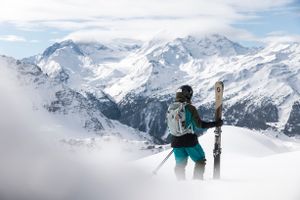 Ein Mann in Skibekleidung und Helm steht im Pulverski mit Scott Freeride Ski in der Hand und Blickt in die verschneite Berglandschaft