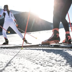 Ein Mann und eine Frau gehen neben der Loipe Langlaufen mit einer Ausrüstung von Kästle in der Skating Variante. Im Hintergrund sind schneebedeckte Berge