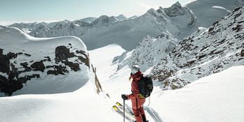Eine Skifahrerin steht am Grat mit Ski im Pulverschnee und blickt in eine winterliche Berglandschaft