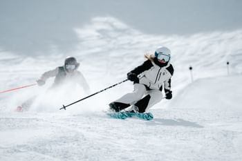 Eine Frau und ein Mann fahren auf einer Piste einen Berg hinunter. Im Hintergrund fährt eine Seilbahn.