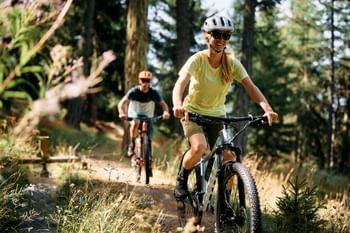 Eine Frau und ein Mann fahren auf ihren E-Fully Mountainbikes mit Helm und Oakley Sonnenbrille einen Schotterweg auf einem Waldtrail entlang