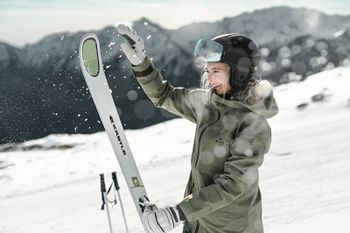 Eine Frau schlägt den Schnee von ihren aufgestellten Skiern auf der Piste in einem Skigebiet mit winterlichen Bergen im Hintergrund 