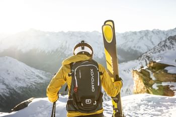 Ein Mann in Skibekleidung und Helm mit Ortovox Rucksack steht mit Kästle Ski am Bergrand und schaut in die winterliche Berglandschaft