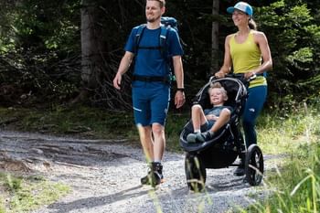 Ein junger Mann und eine junge Frau spazieren im Wanderbekleidung mit Wanderschuhen mit einem Kinderwagen, indem ein Junge gemütlich sitzt, auf einem Waldweg in Serfaus