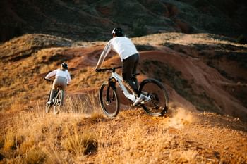 Zwei Fahrradfahrer biken im Sonnenuntergang auf einem Trail am Berggrat. Im Hintergrund sieht man die Berge im goldenen Licht