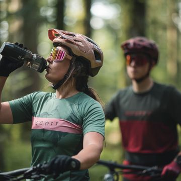 Eine Frau trinkt aus einer Trinkflasche im Hintergrund sitzt ein Mann auf einem Mountainbike und wartet im Wald