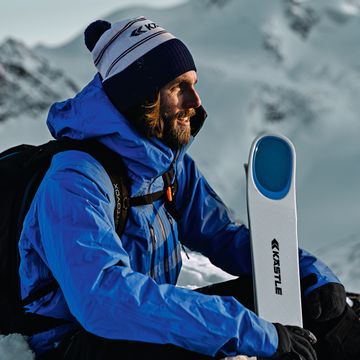 Ein Mann in Skibekleidung und Mütze sitzt im Skigebiet im Schnee und hält seinen Kästle Ski im Arm