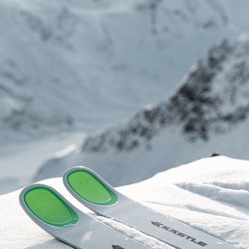 Die Skispitzen der Kästle Ski auf einem Pistenrand im Schnee. Im Hintergrund ist eine winterliche Berglandschaft