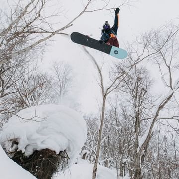 Terje Haakonsen springt im Wald mit einem Burtin Family Snowboard über einen tiefverschneiten Baumstamm in den Tiefschnee