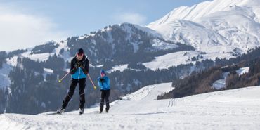 Zwei Personen betreiben Langlauf auf einer präparierten Loipe. Im Hintergrund ist das Skigebiet von Serfaus
