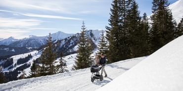 Ein Mann und eine Frau gehen in einer tief verschneiten Winterlandschaft mit einem gemieteten Kinderwagen von Patscheider im Wald spazieren, m Hintergrund ist das Bergpanorama in Serfaus