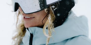 Eine Frau steht in einer weiß verschneiten Berglandschaft. Sie trägt eine hellblaue Jacke, Helm und Schneebrille und blickt lächelnd zur Seite