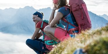 Zwei Frauen ausgestattet mit Ortovox Wanderbekleidung und Rucksäcken machen eine Pause von ihrer Wanderung auf einer Wiese in sommerlicher Berglandschaft
