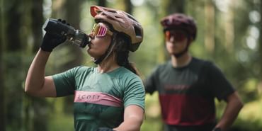 Eine Frau trinkt aus einer Trinkflasche im Hintergrund sitzt ein Mann auf einem Mountainbike und wartet im Wald
