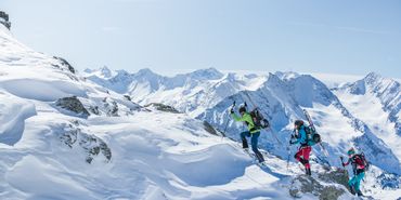 Eine Gruppe von Skifahrern geht eine Tour einen Berggrat hinauf, die Ski sind am Rucksack befestigt im Hintergrund ist eine Bergpanorama mit verschneiten Gipfeln