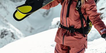 Eine Skifahrerin steht am Grat mit geschulterten Ski und lächelt im Hintergrund ist eine winterliche Berglandschaft