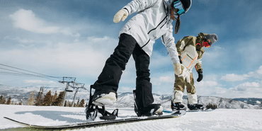 Eine Frau und ein Mann steigen mit ihren Step On Boots in die Burton Step on Bindung auf einer Piste im Skigebiet ein