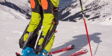 Ein Skifahrer steht auf der Piste mit Againern an den Beinen und Skischuhen befestigt. Im Hintergrund ist ein Bergpanorama 