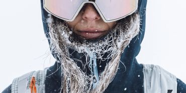 Eine Frau steht mit Skibekleidung, Helm, und Schneebrille frontal. Ihre Haare sind von der Kälte vereist und sie blickt in die Kamera