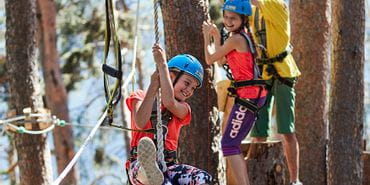 Lächelndes Mädchen schaukelt auf einer Boje im Kletterseilpark X-Trees. Im Hintergrund lachen auf einer Plattform Kinder.