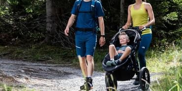 Ein junger Mann und eine junge Frau spazieren im Wanderbekleidung mit Wanderschuhen mit einem Kinderwagen, indem ein Junge gemütlich sitzt, auf einem Waldweg in Serfaus
