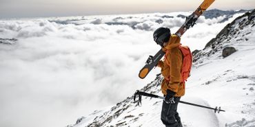 Ein Mann steht an einem Grat in wolkenverhangenen Bergen und hat die Ski auf den Schultern