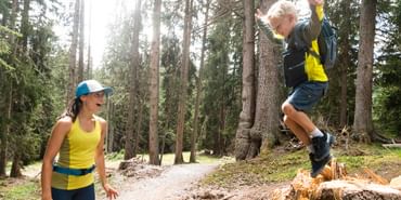 In einem Wald springt ein Junge von einem abgesägten Holzstamm. Daneben steht eine junge Frau und lächelt