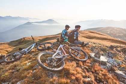 Zwei Männer machen von ihrer Bike Tour eine Pause und sitzen auf einem Hügel in einer sommerlichen Berglandschaft in Serfaus ihre Mountainbikes sind ins Gras gelegt