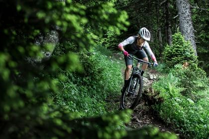 Eine Frau fährt auf einem Fully MTB von Scott einen Trail durch den Wald über Baumwurzeln hinunter