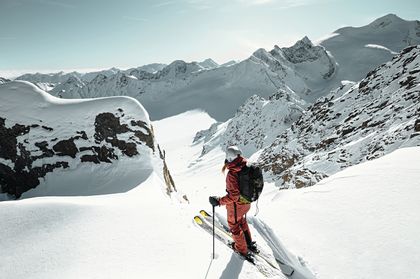 Eine Skifahrerin steht am Grat mit Ski im Pulverschnee und blickt in eine winterliche Berglandschaft