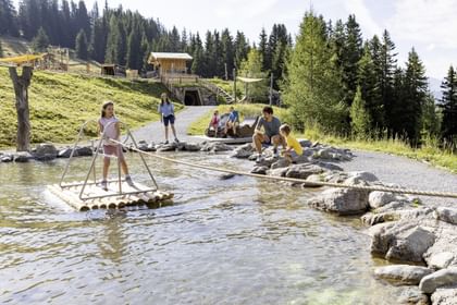 Kinder spielen im Murmliwasser und ein Mädchen steht auf einem Floß und zieht sich über ein Seil auf die andere Seite des Flusses