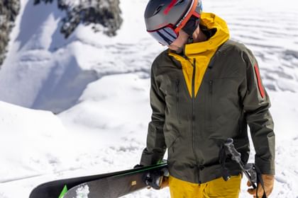 Ein Mann läuft am Grat eines verschneiten Gipfels entlang und trägt seine Ski in der einen einen, die Stöcke in der anderen Hand